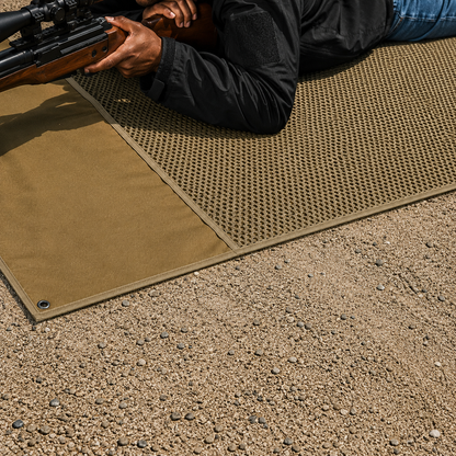 Close-up view of a person lying prone on the tan shooting mat holding a rifle, demonstrating how the padded surface protects the shooter from rough gravel and dirt terrain.